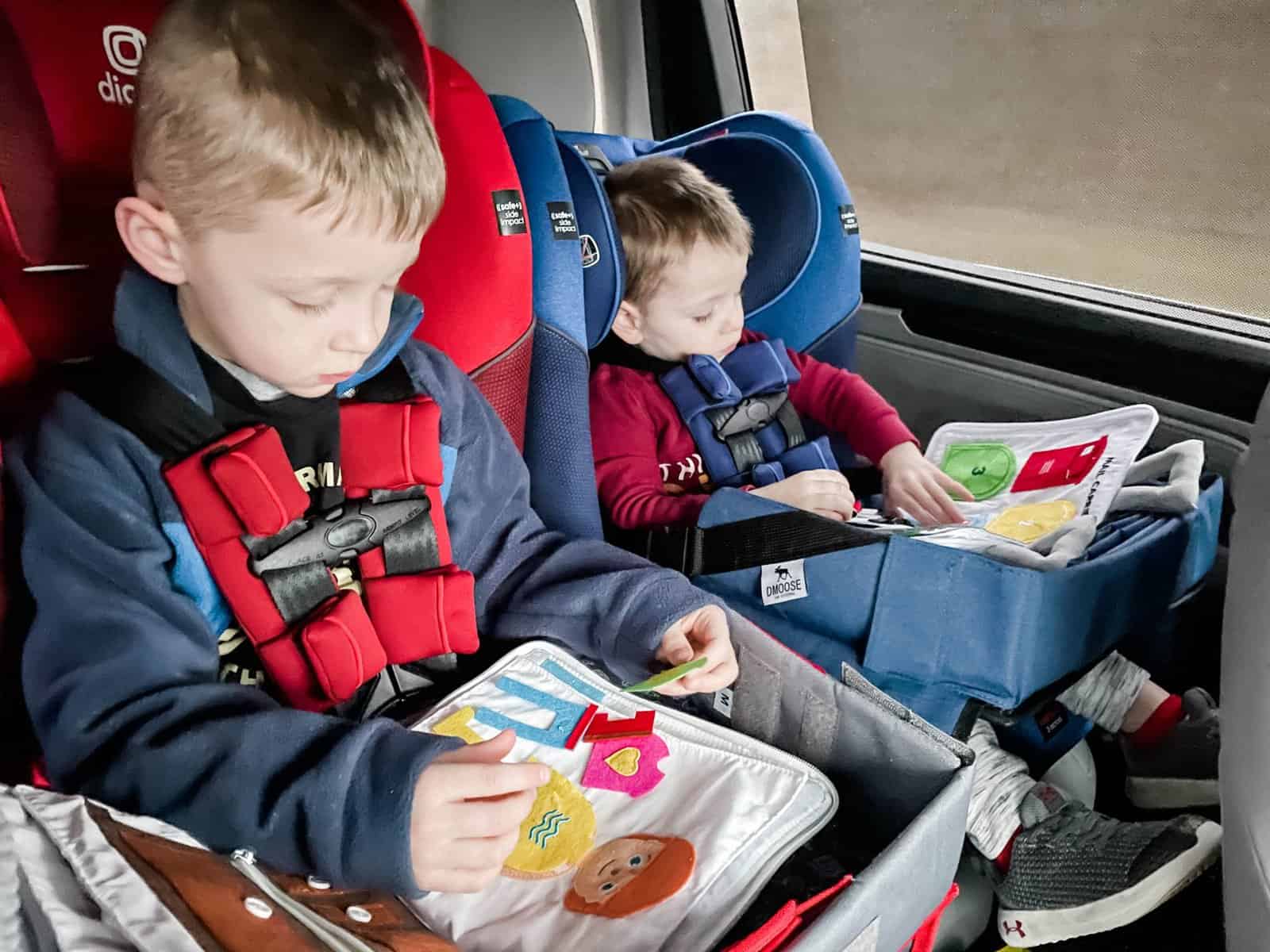 Two boys in car seats while driving with kids on a long road trip playing with items from their travel activity pack for kids